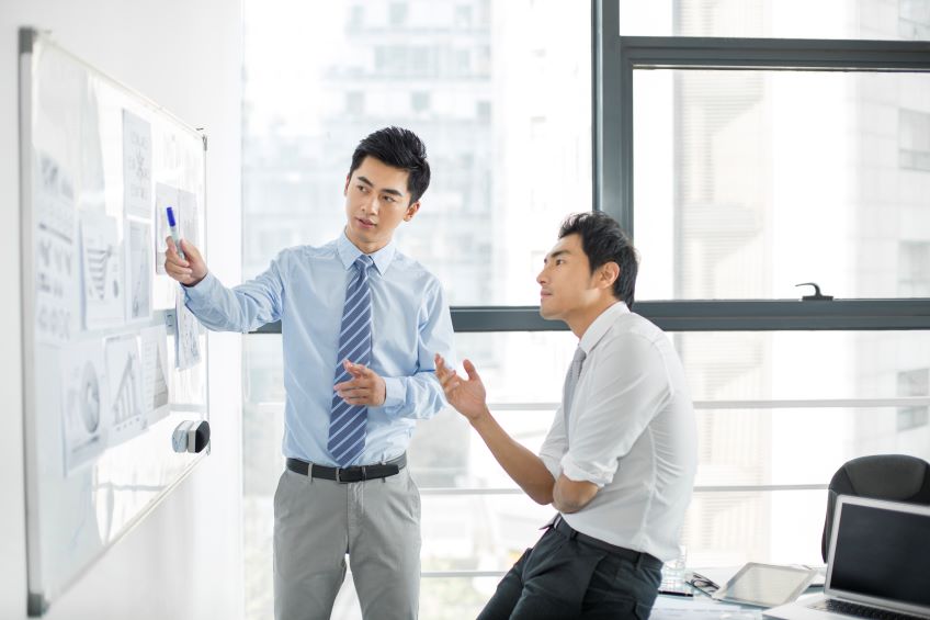 A person points at a document posted on a white board as another individual watches and gestures