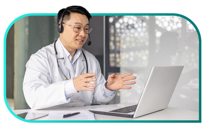 Person wearing a white physician’s coat and a headset sitting at a desk talking on a laptop.